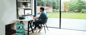 Woman sat at desk at home looking at her notepad pad and laptop with printer, plants and a chair in the foreground